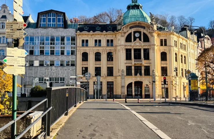 Панорама улицы в Карловых Варах с видом на красивое светло-желтое здание в стиле модерн с медным куполом. На переднем плане пешеходный мост с черными перилами и указатель с табличками