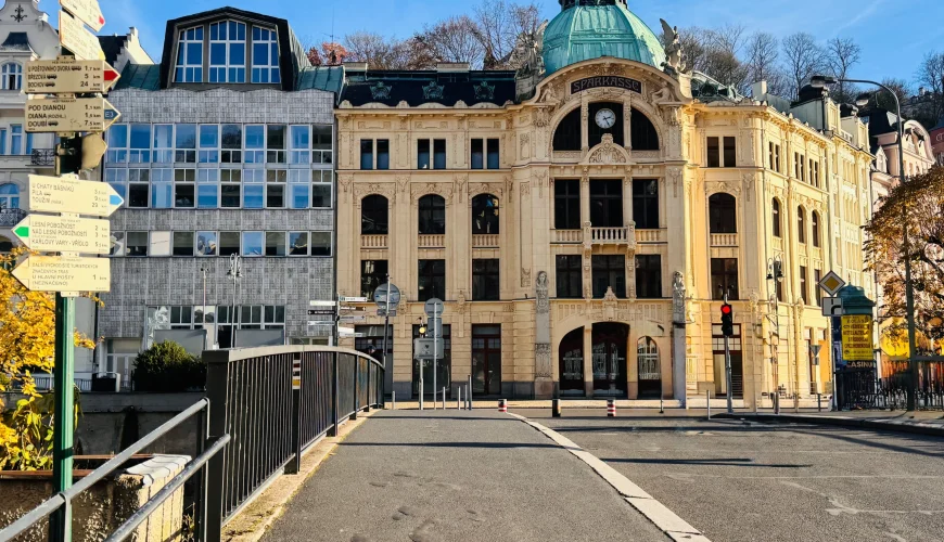Панорама улицы в Карловых Варах с видом на красивое светло-желтое здание в стиле модерн с медным куполом. На переднем плане пешеходный мост с черными перилами и указатель с табличками