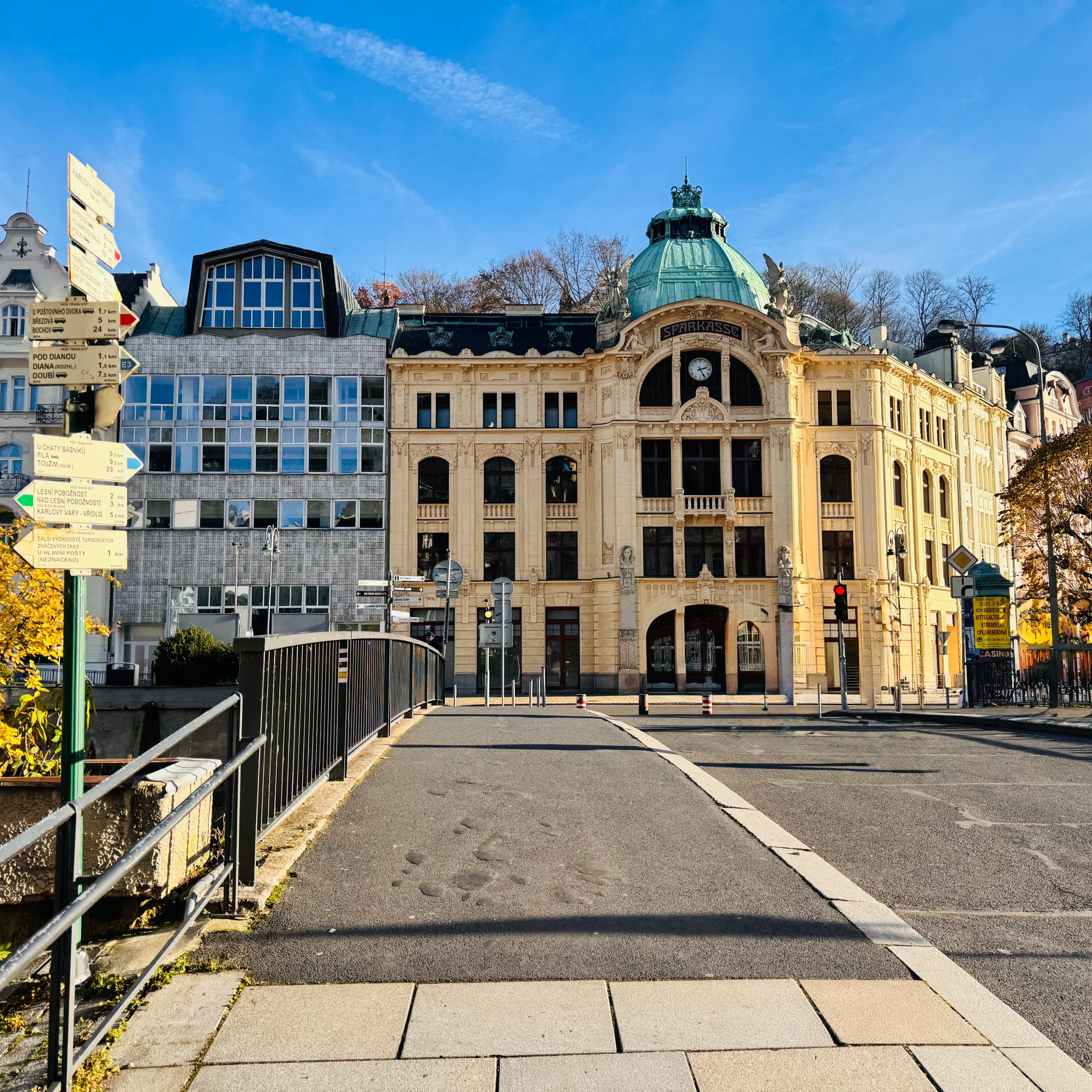 Панорама улицы в Карловых Варах с видом на красивое светло-желтое здание в стиле модерн с медным куполом. На переднем плане пешеходный мост с черными перилами и указатель с табличками